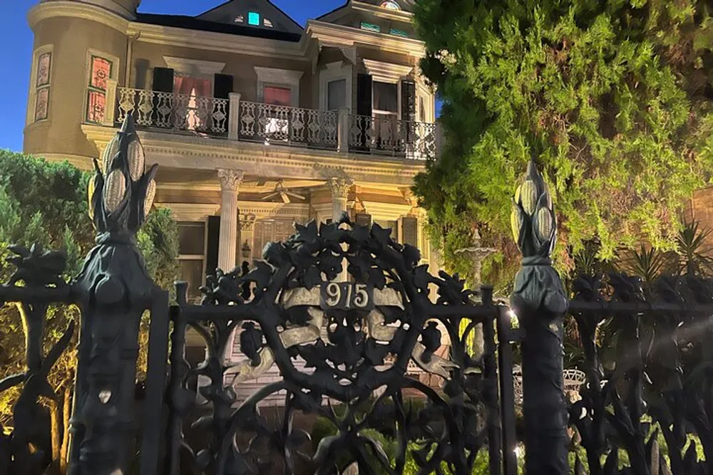 The image shows an elegant two-story historical house viewed through an ornate wrought-iron gate with the number 915 on it set against a twilight sky