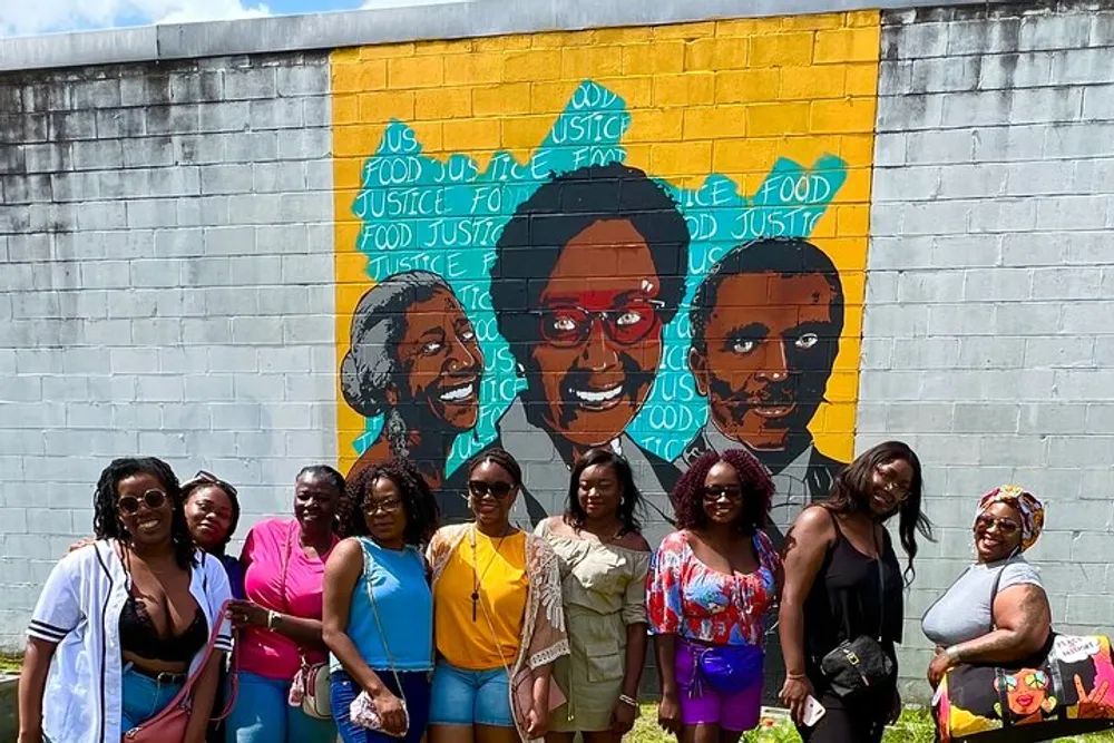 A group of people is posing in front of a colorful mural dedicated to food justice featuring portraits and repeated slogans advocating for the cause