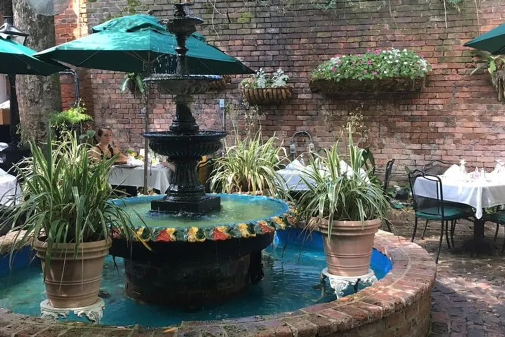 The image shows a charming outdoor restaurant courtyard with a multi-tiered fountain surrounded by greenery and tables with a person seated in the background under an umbrella