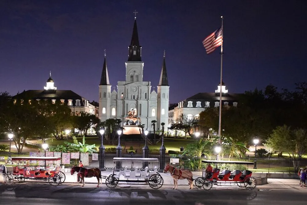 The image shows a night view of Jackson Square with the Saint Louis Cathedral lit up in the background horse-drawn carriages waiting at the front and the American flag waving to the side