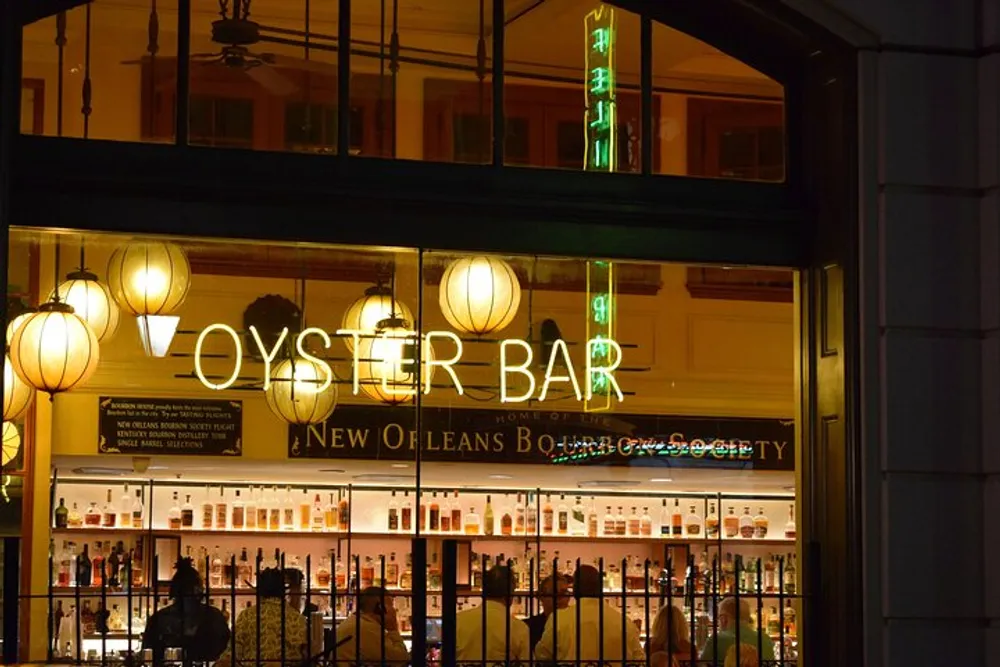 The image shows the warmly lit interior of an Oyster Bar with neon signs spherical lamps and shelves stocked with bottles visible through large glass windows from the street at nighttime