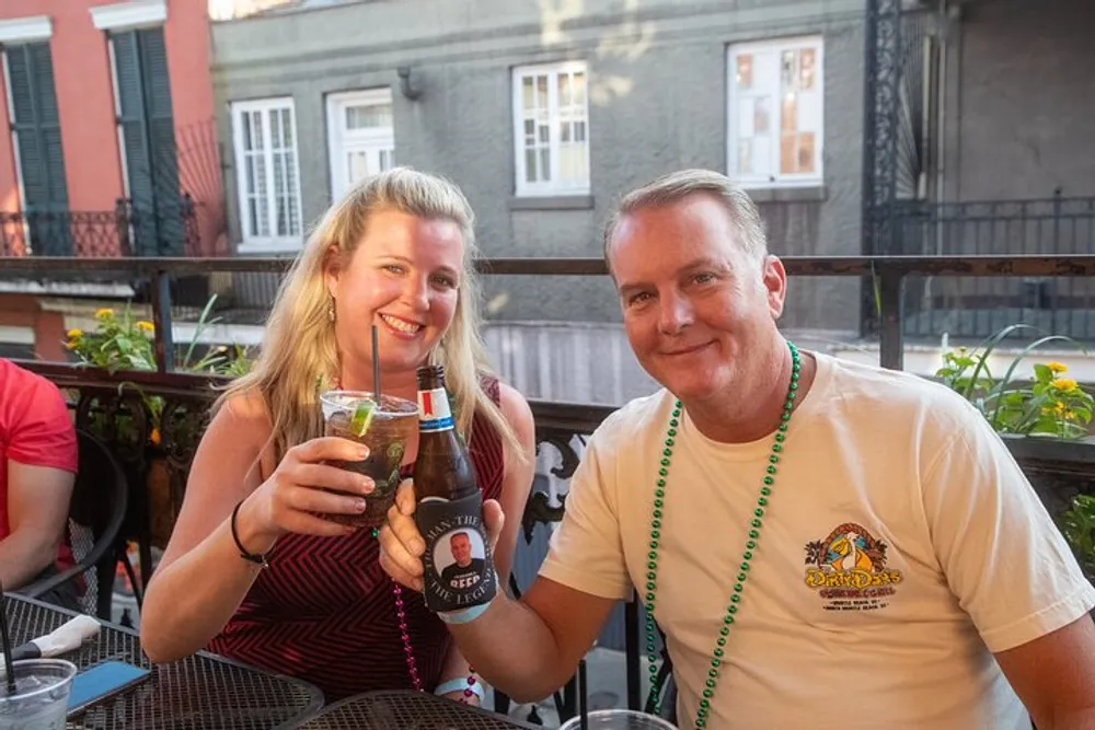A woman and a man are smiling and enjoying drinks on a balcony with festive beads around their necks