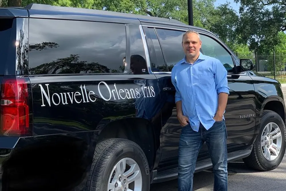 A man is standing and smiling next to a large black SUV that has Nouvelle Orleans written on its side
