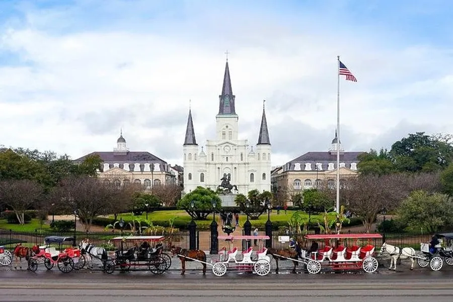 Horse-drawn carriages wait in front of the iconic Saint Louis Cathedral in Jackson Square, New Orleans, under a cloudy sky with the American flag flying to the right.