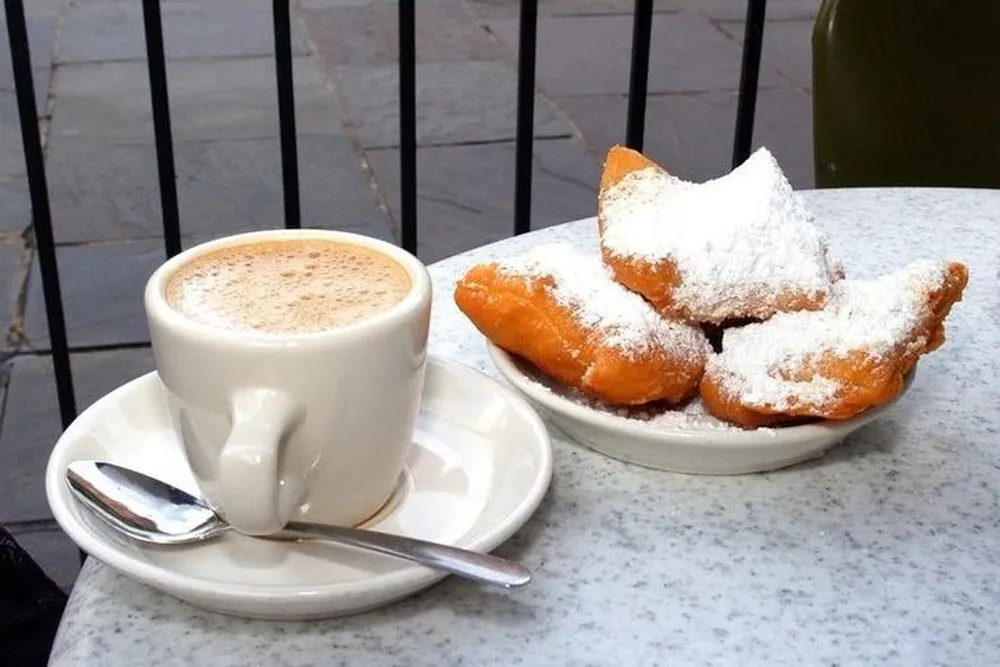 A cup of coffee and a plate of beignets dusted with powdered sugar are sitting on a marble table presumably at an outdoor cafe