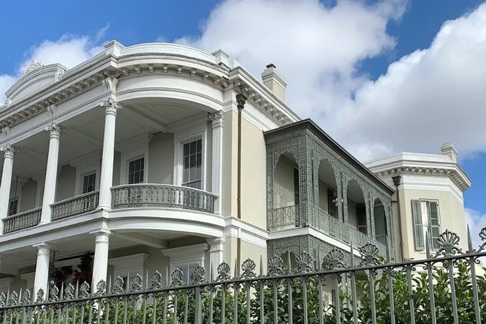 The image shows a grand two-story white house with ornate iron balconies and railings set against a partly cloudy sky