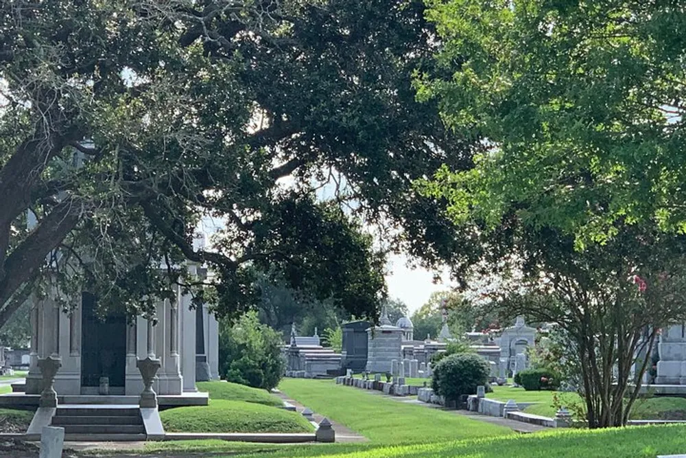 The image shows a peaceful and well-maintained cemetery dotted with various mausoleums and headstones under the shade of large sprawling trees
