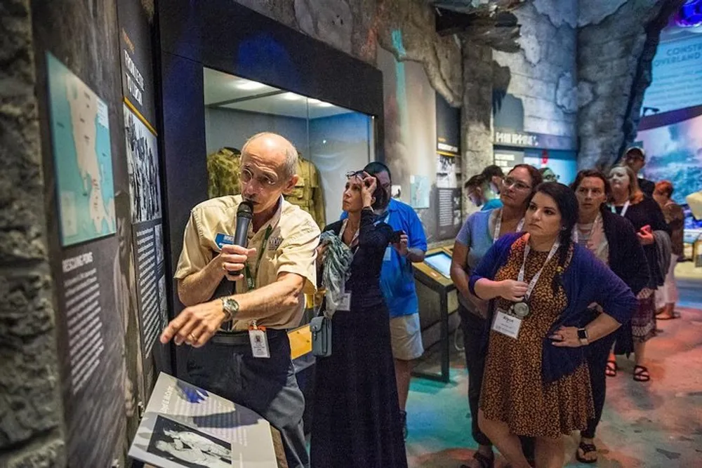 A guide is speaking into a microphone while giving a presentation to a group of attentive visitors inside a museum or exhibition