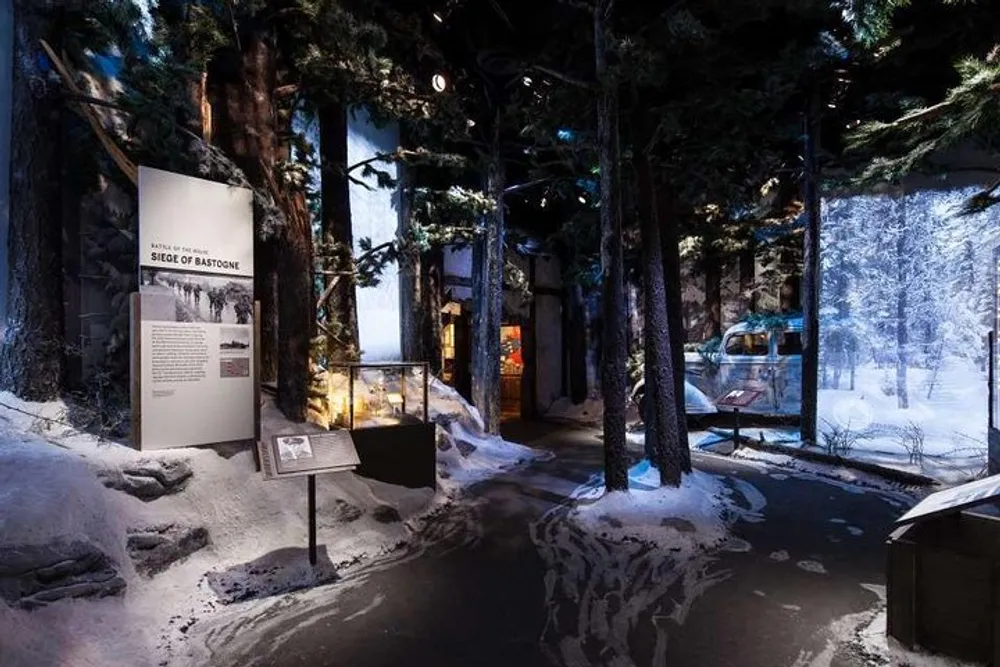 The image depicts an immersive museum exhibit with a snowy forest setting featuring informational stands a backdrop with a wintery scene and a military vehicle to represent the historical context of the Siege of Bastogne
