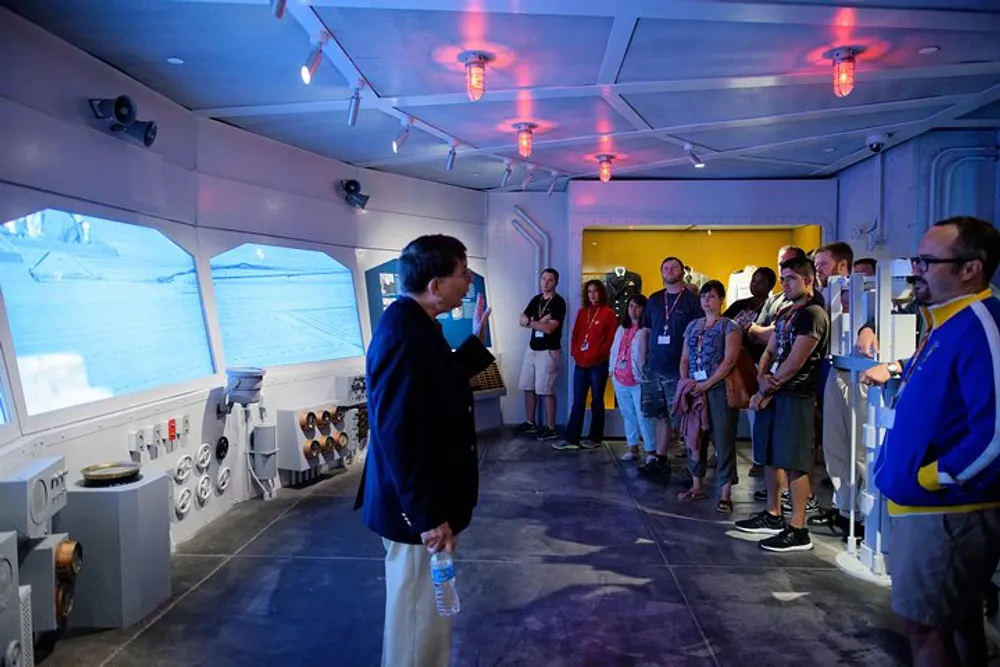 A group of people is listening to a presenter inside a room that simulates the interior of a ship or submarine with a view of icy waters projected on one wall
