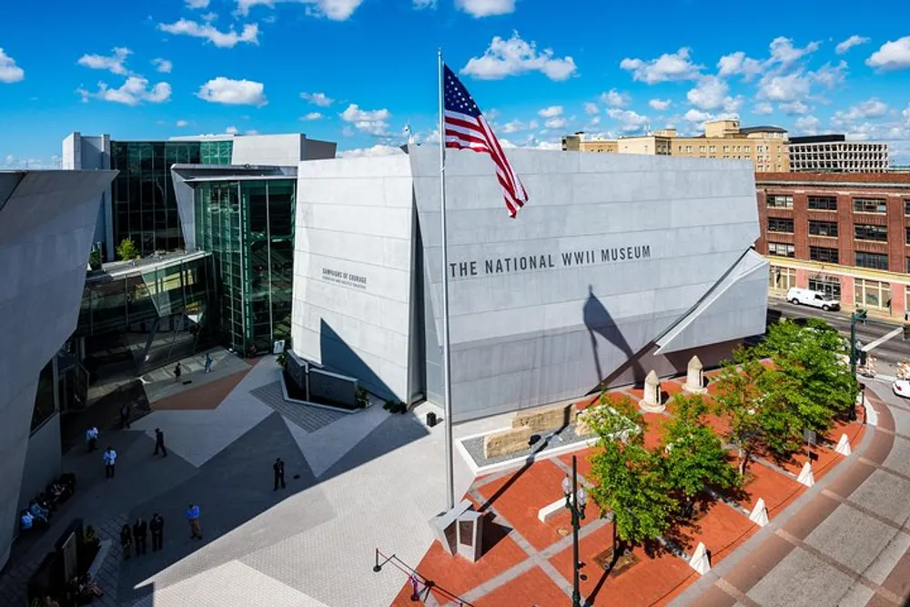 The image shows a sunny day view of the exterior of The National WWII Museum with an American flag flying in front and several people walking around the entrance plaza