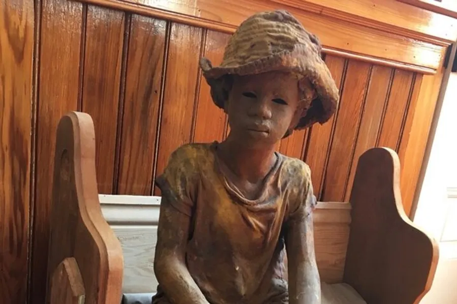 The image shows a life-like sculpture of a child sitting on a wooden bench, adorned with an oversized floppy hat.