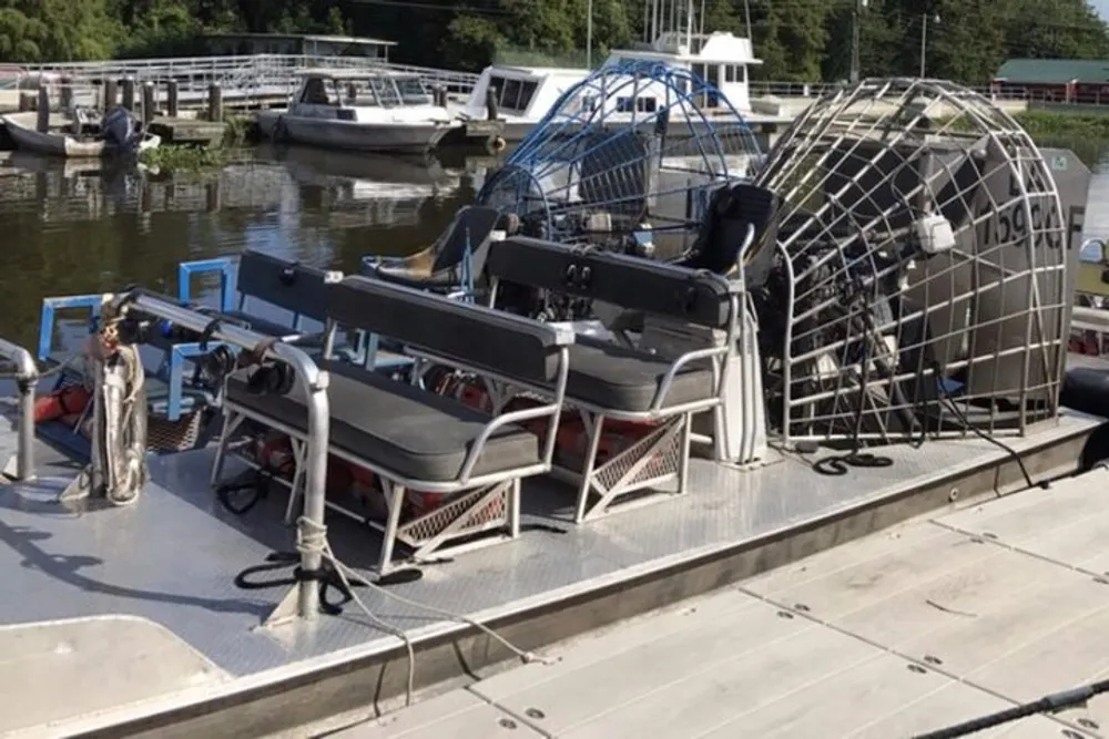 The image shows a large airboat with seating and protective caging docked at a marina alongside other boats