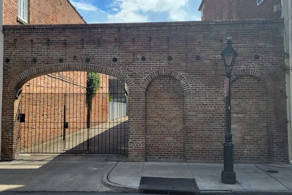 The photo features a brick archway with a metal gate partially open and an antique-style streetlamp standing in the foreground on a paved street