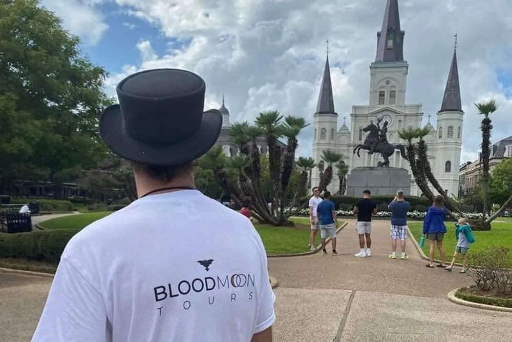 A person wearing a top hat and a T-shirt that says Bloodmoon Tours is standing with their back to the camera looking towards a group of people and a historic church with white steeples in the background