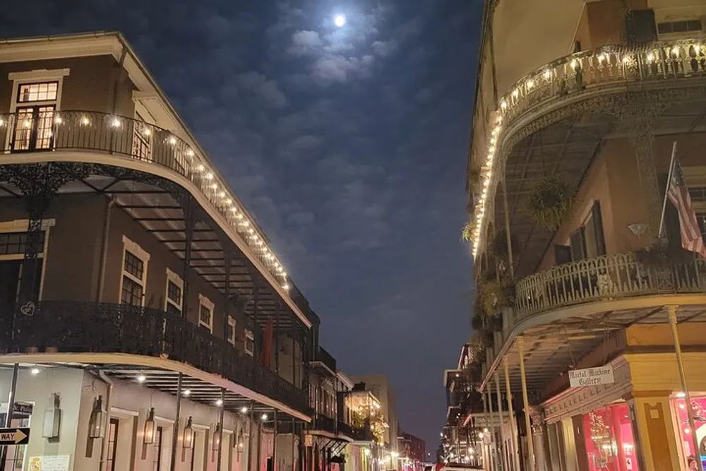 An evening view of a charming street lined with historic buildings featuring ornate balconies illuminated by warm lights under a twilight sky with a visible moon