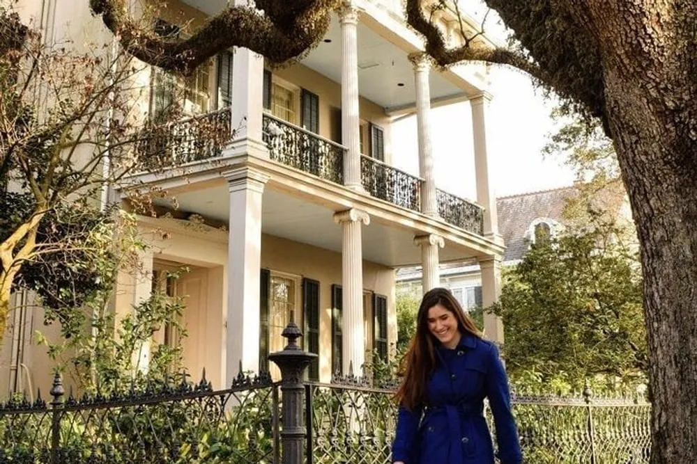 A smiling person in a blue coat stands in front of a large house with a balcony and wrought-iron fence framed by mature trees