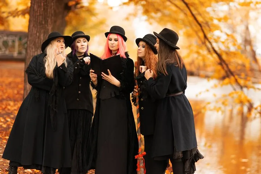 Five people dressed as witches are gathered around a book, engaging in a lighthearted moment amidst an autumnal setting.