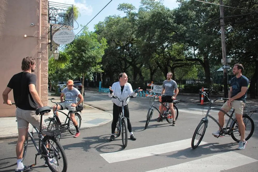 A group of men on bicycles are pausing at an intersection engaging in a conversation on a sunny day with lush trees and a clear sky in the background