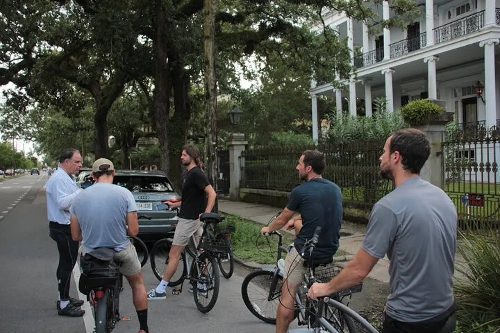 A group of cyclists is having a conversation with a person who is standing next to a car on a street lined with trees and historic homes