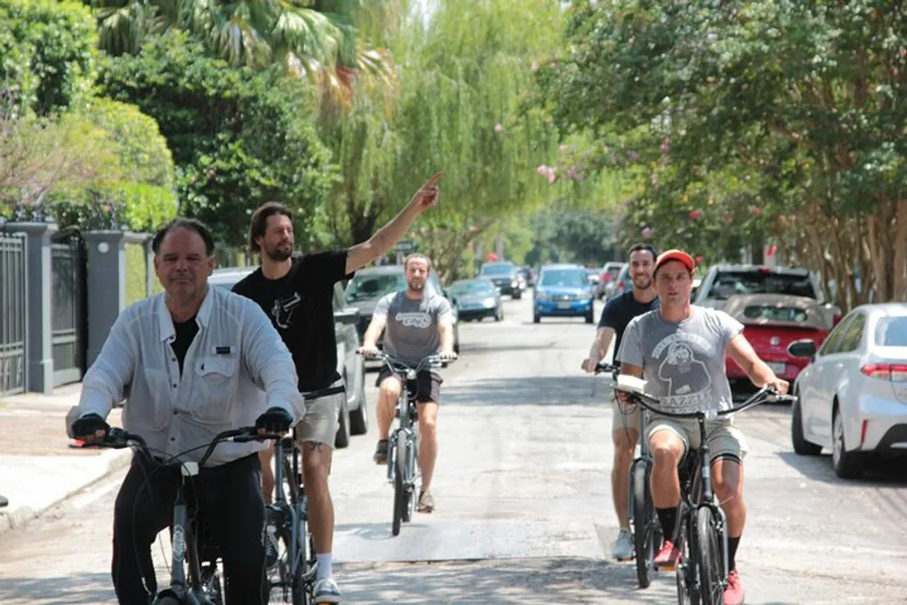 A group of men are casually riding bikes down a tree-lined street with one of them pointing to something in the distance