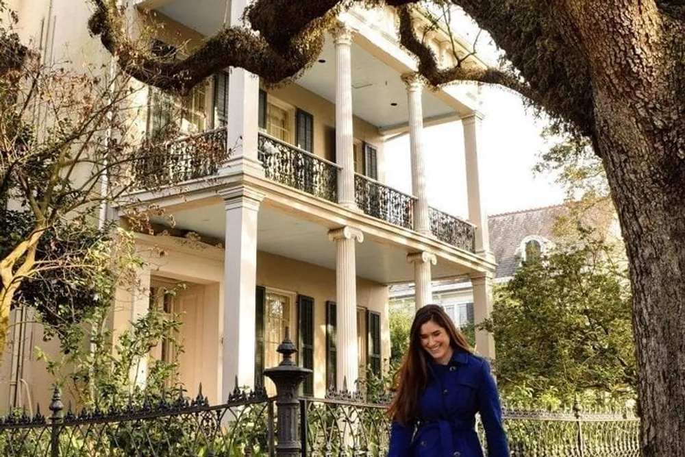 A woman in a blue coat is smiling in front of a grand two-story house with columns and balconies shaded by an old tree with sprawling branches