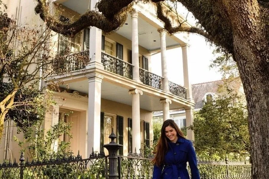 A woman in a blue coat is smiling in front of a grand, two-story house with columns and balconies, shaded by an old tree with sprawling branches.