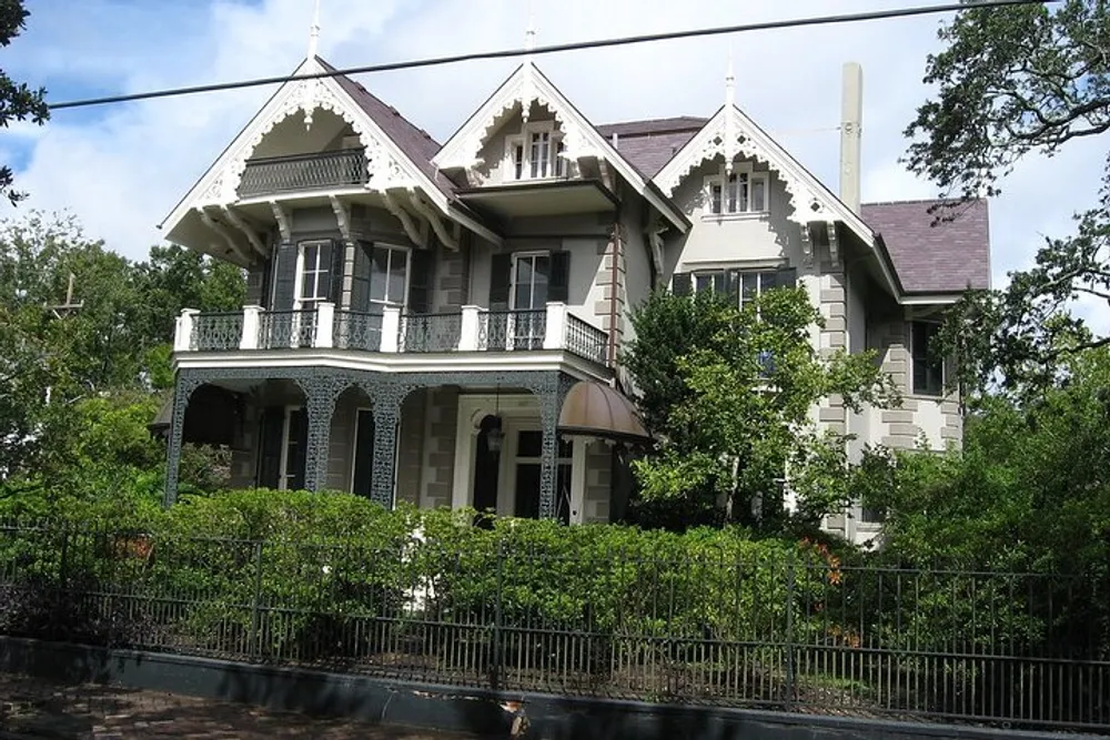 The photo shows a grand Victorian-style house with detailed trim a second-story balcony and a lush garden fronting a wrought iron fence