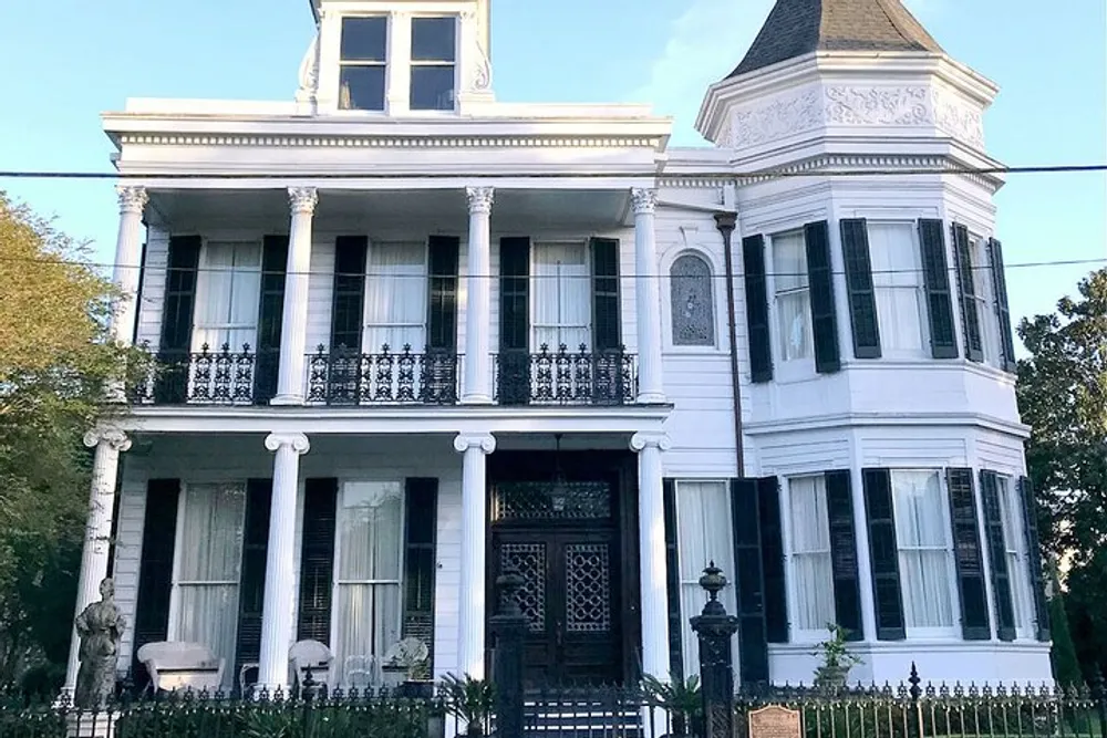The image shows a stately white two-story house with a grand entrance intricate wrought iron balconies and a distinctive tower-like structure on the roof evoking the architectural style commonly found in historic Southern US neighborhoods