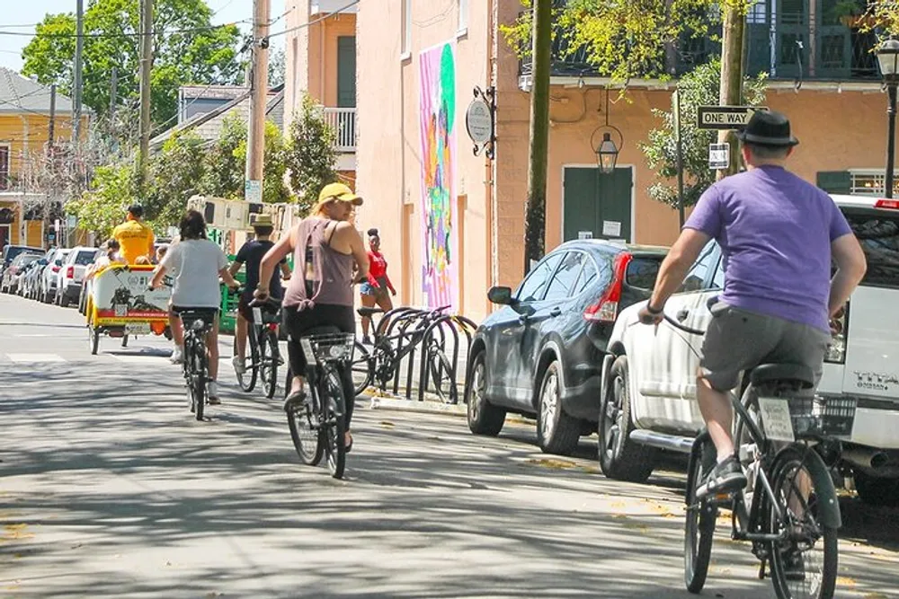 The image shows a group of people cycling down a sunlit street lined with parked cars and adorned with a colorful mural on the building to the left