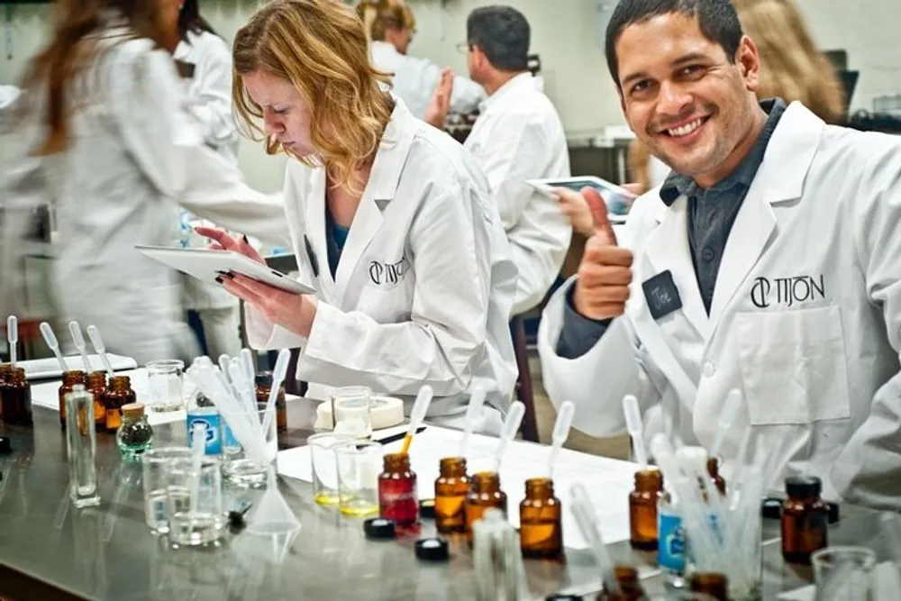 In a laboratory setting with various bottles and lab equipment on the table a man in a white lab coat smiles and gives a thumbs-up while a woman next to him is focused on her work examining something on a tablet