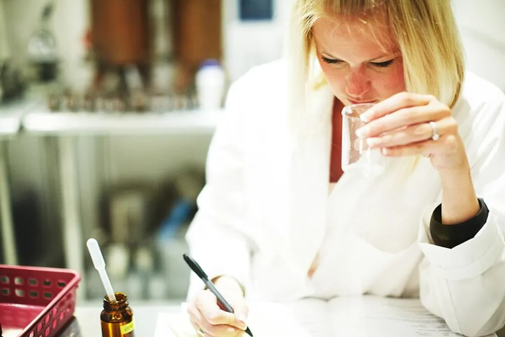 A scientist in a lab coat is intently smelling a substance in a beaker while making notes