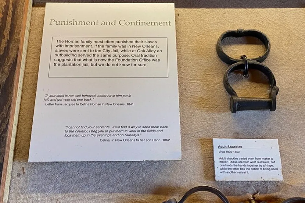 The image depicts a museum exhibit detailing the punishment and confinement of slaves with an informational sign and a pair of adult shackles from around 1830-1860