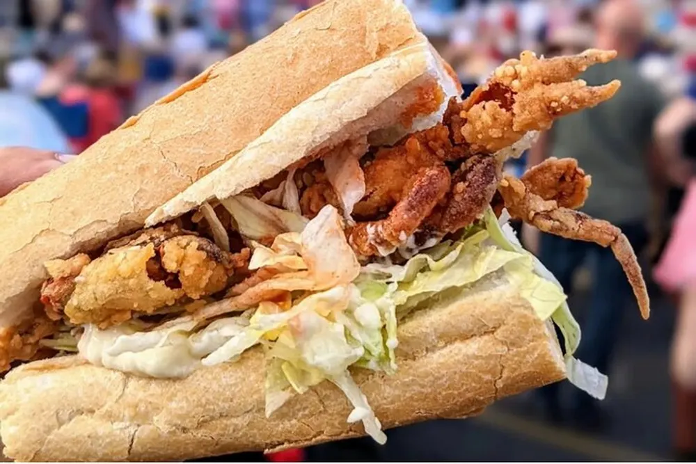 A hand is holding a po boy sandwich filled with fried seafood and lettuce with a blurred crowd in the background