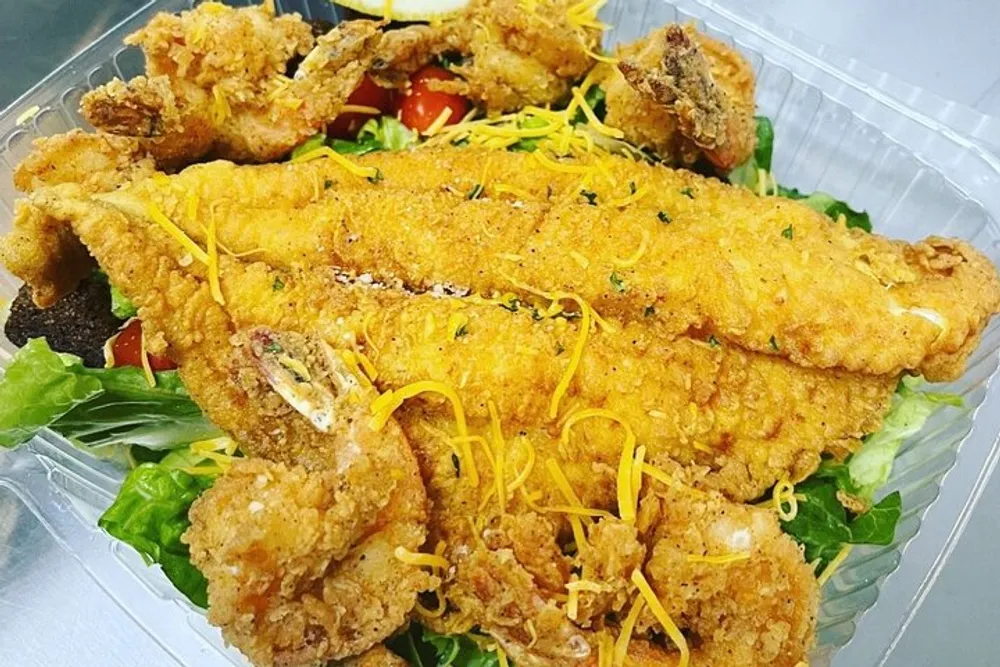 The image shows a salad topped with crispy fried seafood likely catfish and shrimp garnished with shredded cheese and herbs served in a clear plastic takeout container