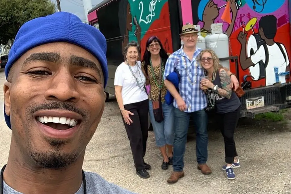A person is taking a selfie with a group of four cheerful people standing in the background near a colorful vehicle adorned with cartoon-like art