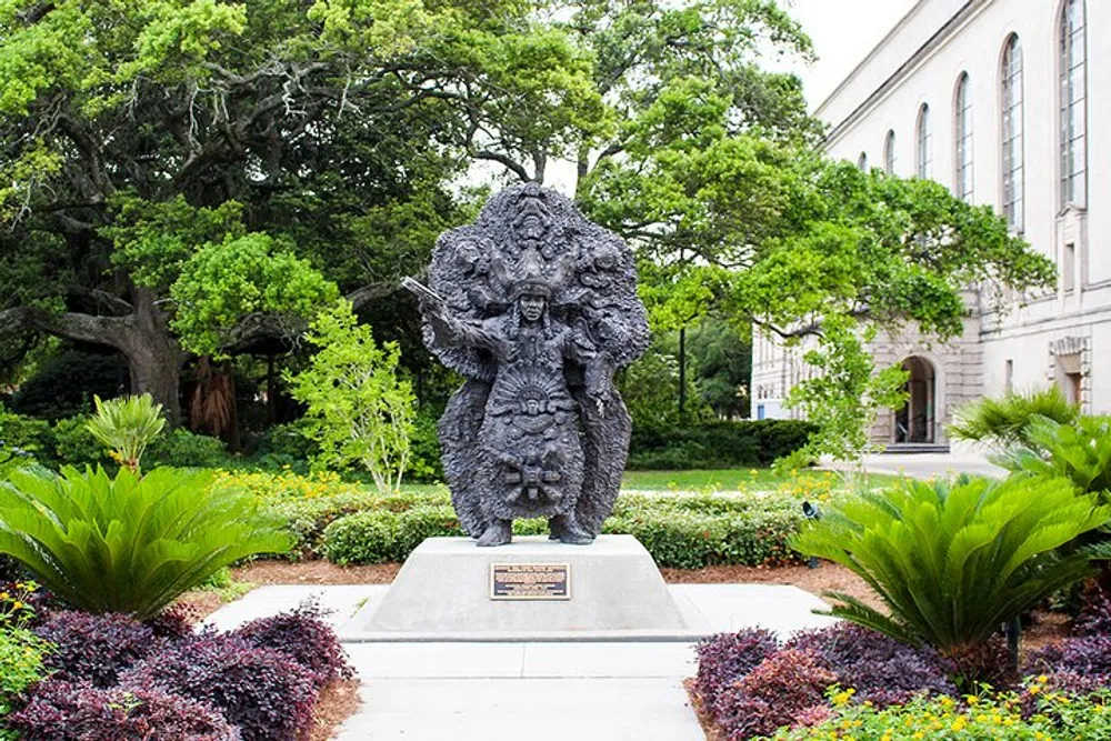 The image shows a statue situated in a landscaped area featuring a figure with multiple faces and textured details set against a backdrop of trees and a building