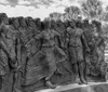 The image shows a black and white photograph of a detailed expressive sculpture depicting a group of figures possibly portraying a historical or significant cultural event