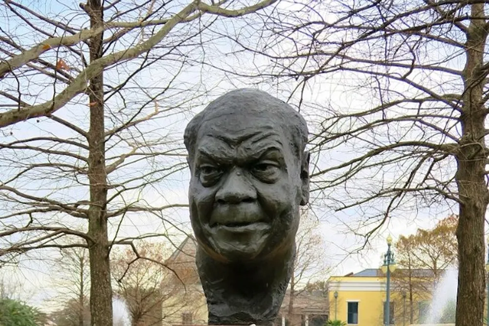 The image shows a large outdoor sculpture of a human head with an exaggerated expression set against a background of leafless trees and a cloudy sky