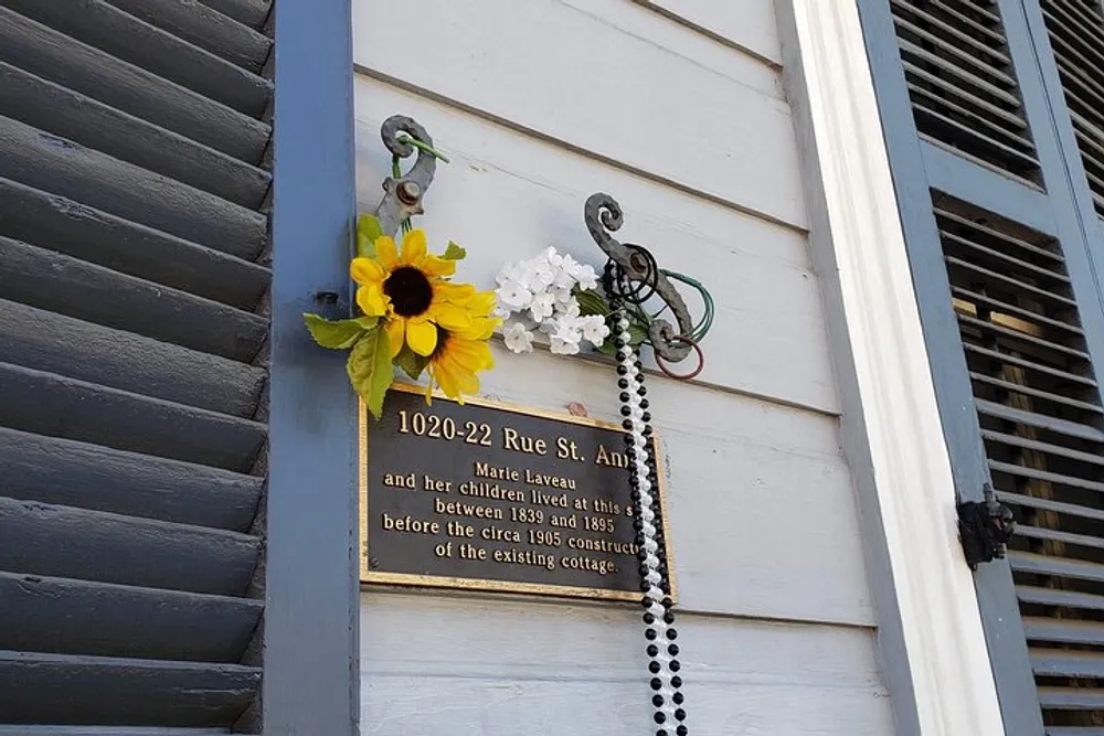 A decorative plaque affixed to a wooden wall with artificial flowers and beaded necklaces commemorates Marie Laveaus residence at this location from 1836 to 1885 before the existing cottage was constructed