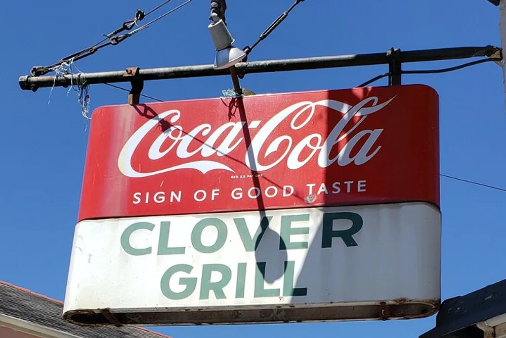 The image shows a vintage Coca-Cola sign above the Clover Grill sign both mounted on a metal structure against a clear blue sky