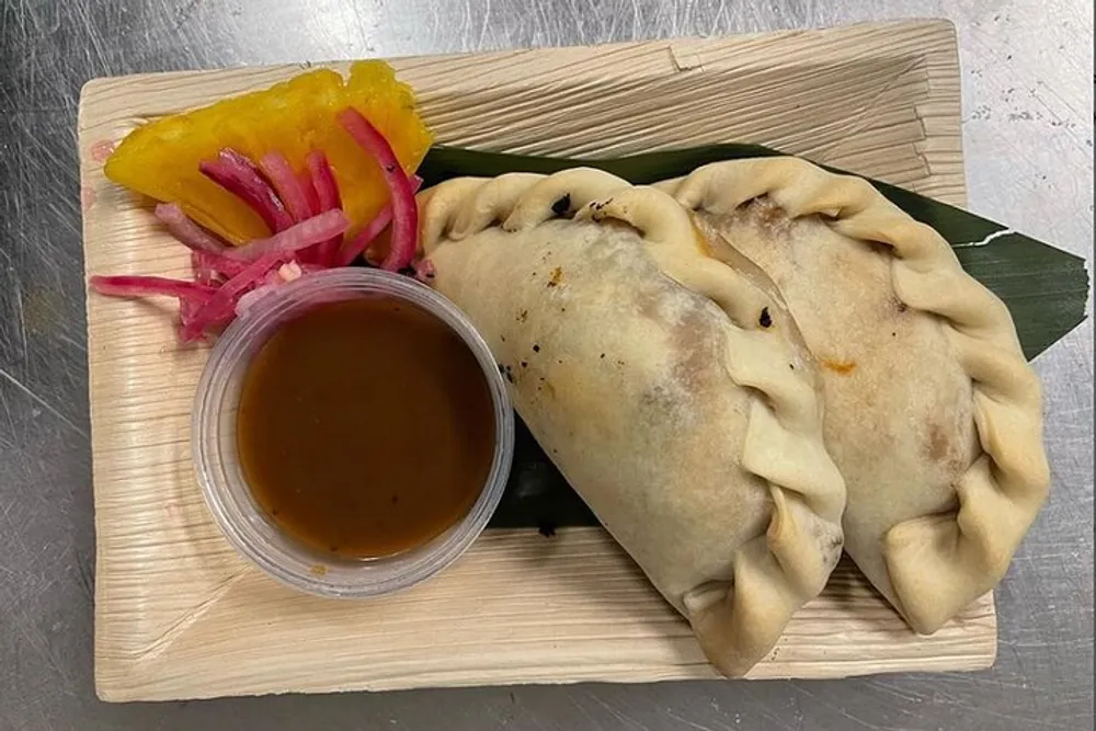 The image shows two empanadas on a wooden plate accompanied by pickled onions a slice of pepper and a cup of sauce