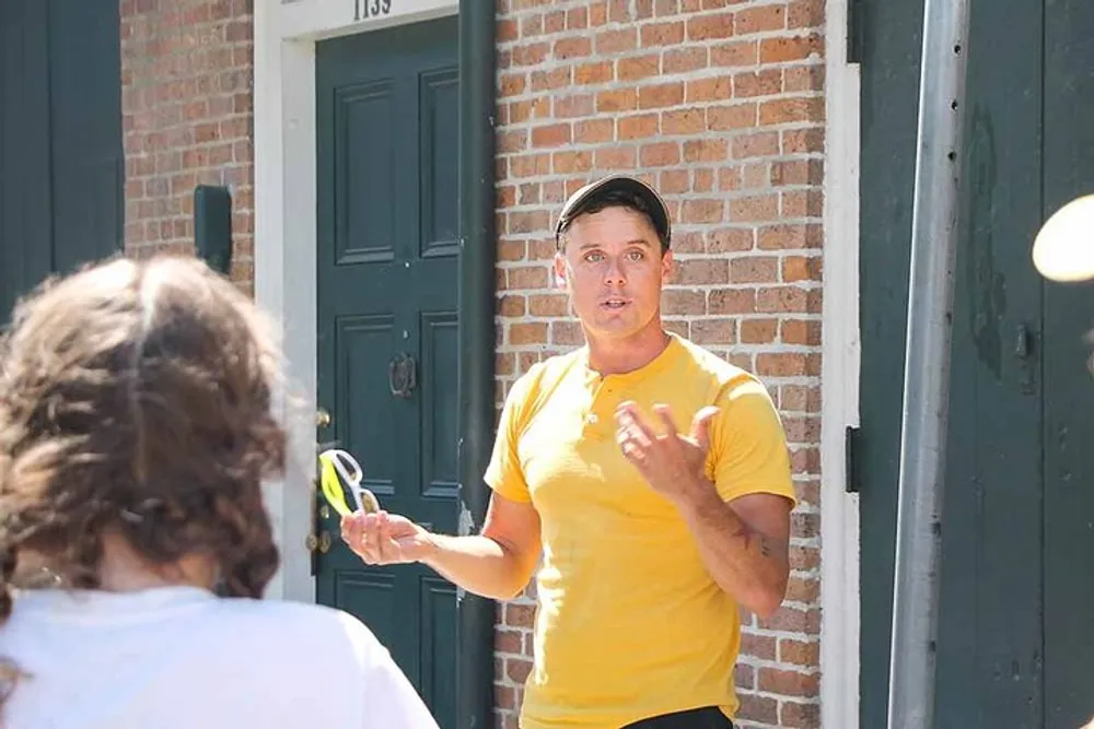A person in a yellow t-shirt and a cap is talking and gesturing with their hands while holding sunglasses with another persons back to the camera in the foreground and a brick building with a dark green door in the background