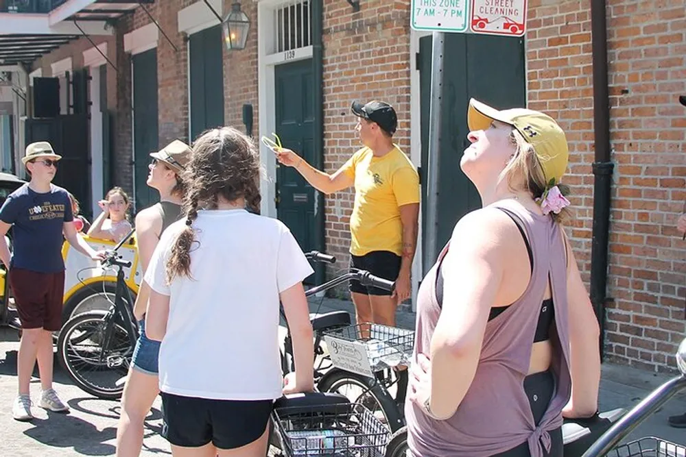 A group of people some on bicycles is attentively listening to a person who appears to be a guide while a woman in a cap looks up towards the sky or a building