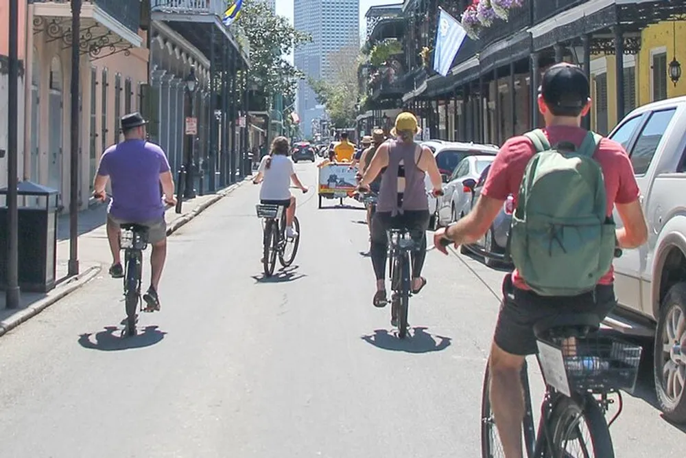 Cyclists are riding down a sunny urban street lined with parked cars and buildings with balconies