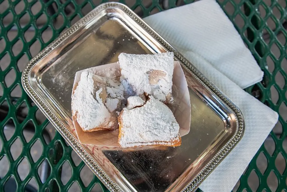 Powdered sugar-dusted pastries are served on a metal tray over a paper napkin and a green mesh table