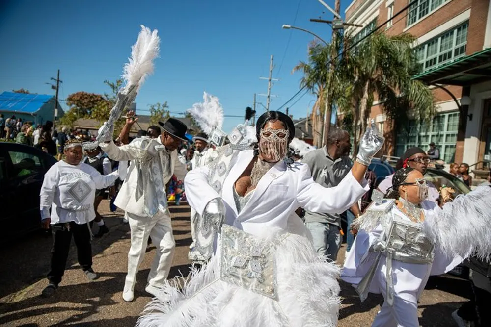 A group of people dressed in elaborate white and silver costumes with feathers and masks are participating in a vibrant street parade