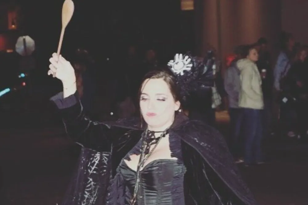 A person is dressed in a dark gothic-inspired costume playfully holding up a wooden spoon with their eyes closed and a whimsical expression on their face possibly during a night-time outdoor event
