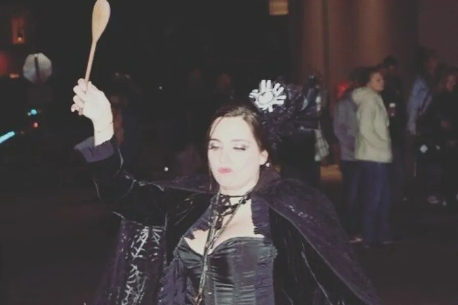 A person is dressed in a dark, gothic-inspired costume, playfully holding up a wooden spoon, with their eyes closed and a whimsical expression on their face, possibly during a night-time outdoor event.