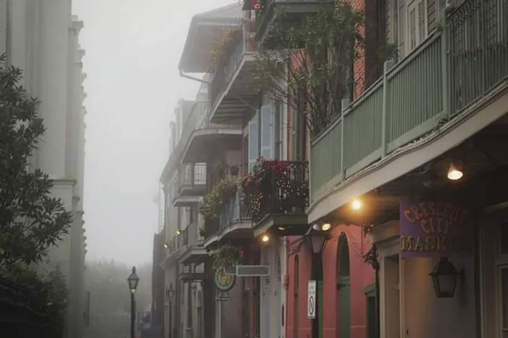 The image displays a misty view of a street lined with quaint colorful buildings and balconies adorned with plants with an old-fashioned lamppost in the foreground and a sign for Crescent City Market